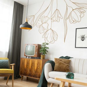 May include: A living room scene with a vintage wooden cabinet, a retro television, and a white sofa. A large floral wall decal in gold lines the wall. A framed bee print hangs on the wall. A green throw blanket and a yellow chair add color to the room.