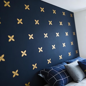 May include: A dark navy blue wall featuring a repeating pattern of small, gold-coloured four-petaled flower decals. Below the wall, a bed is visible with a navy blue pillow featuring a white grid pattern and a solid light grey pillow.