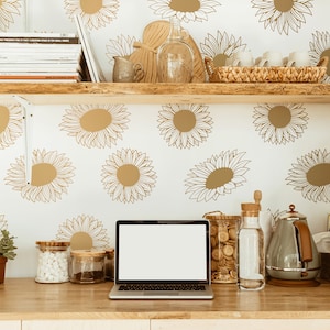 May include: A laptop computer with a blank screen sits on a wooden countertop in front of a white wall with a sunflower pattern. There are various kitchen items on the counter, including a glass jar with a cork lid, a glass bottle with a cork lid, a gray electric kettle, and a wooden box with the text "Olive Tea" printed on it.