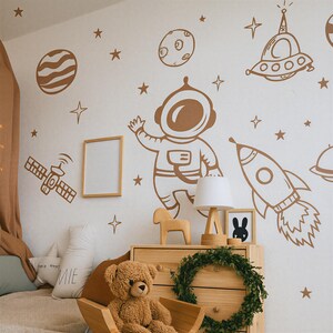 May include: A child's bedroom wall decorated with brown space-themed decals, including an astronaut, rocket, planets, stars, and a satellite. A wooden dresser with a teddy bear, lamp, and framed art is in front of the wall.