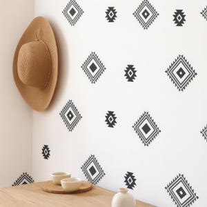 May include: A beige straw hat hangs on a white wall adorned with black geometric patterns. A wooden table holds two small white bowls and a cork coaster. A small white vase sits on the table.