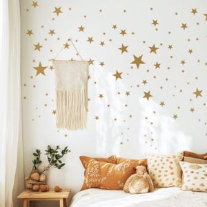 May include: A white wall adorned with gold star decals of varying sizes. A woven wall hanging with fringe hangs from the wall. Pillows in shades of orange and cream rest on a bed. A small wooden table holds a plant and decorative items.