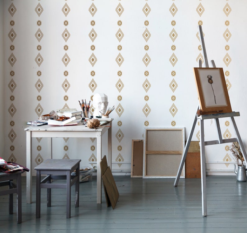 May include: A white room with a wooden table and two stools. The walls are decorated with a gold geometric pattern wallpaper. There is an easel with a blank canvas and a painting on it.