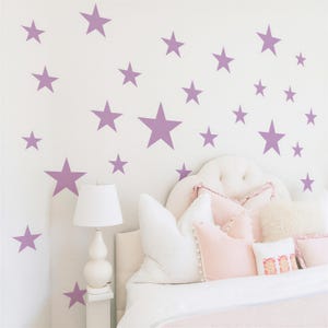 May include: A bedroom scene featuring a white bed with a tufted headboard and various pillows in shades of white and pink. Lavender star decals are affixed to the white wall above the bed, creating a whimsical and decorative theme.