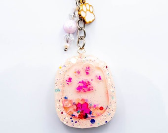 Peut inclure: Un porte-clés en résine rose et blanche avec une empreinte de patte incrustée de paillettes. Le porte-clés a un anneau de clé argenté et un petit charme en forme d'empreinte de patte blanche.