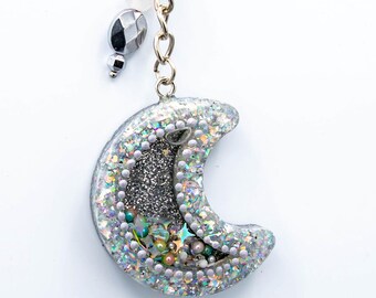 Può includere: Un portachiavi a forma di mezzaluna argentata con anello e catena color oro. La luna è ricoperta di glitter e piccole perline. Il centro della luna è riempito di glitter argentati. Il portachiavi ha un piccolo accento di perline argentate e bianche.