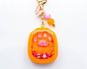 Peut inclure: Porte-clés en résine orange avec une empreinte de patte rose et des paillettes à l'intérieur. Le porte-clés est doté d'une chaîne dorée et de perles roses et orange.