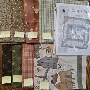 May include: Assortment of fabric pieces with various patterns, including holly, polka dots, snowflakes, and snowmen. A pattern sheet titled "Too Many Men" is also present. The fabrics are in shades of brown, green, and cream.