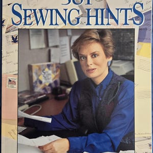 May include: A book cover with the title "501 Sewing Hints" from the viewers of "Sewing with Nancy". The cover features a woman in a blue shirt and a black vest, sitting at a desk with papers and envelopes.