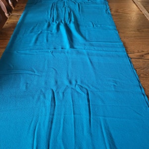 May include: A long, rectangular piece of teal fabric with a textured surface. The fabric is laid out on a wooden surface, with the teal color contrasting against the brown wood.