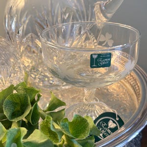 May include: A clear glass champagne coupe with shamrock designs, resting on a silver tray with green foliage. A matching crystal pitcher is in the background. The coupe has a green label that reads "Lamon Glass".