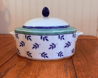 May include: A white ceramic lidded tureen featuring a repeating pattern of small dark blue leaves. The lid has a dark blue knob and a green border. The tureen has two side handles and a scalloped edge design, providing a classic look for serving.