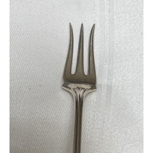 May include: A silver fork with three tines. The fork has a decorative design where the tines meet the handle. The handle is long and slender. The fork is made of a shiny metal.