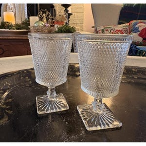 May include: Two clear, vintage-style drinking glasses. Each glass features a diamond-patterned exterior and a square base with a starburst design. The glasses are displayed on a dark, metallic tray.