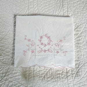 May include: White linen pillowcase with pink floral embroidery. The embroidery features a floral wreath and delicate vines. The pillowcase has a scalloped edge, adding a touch of elegance to the design.