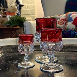 May include: Three clear glass goblets with ruby red rims. The lower section of the goblets has a detailed, patterned design. The goblets are arranged on a dark, reflective surface, highlighting their elegant form and colour contrast.