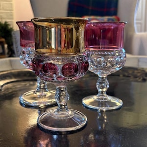 May include: Three decorative wine glasses with gold rims and ruby red bowls. The glasses have a clear, textured stem and base. The glasses are arranged on a reflective surface.