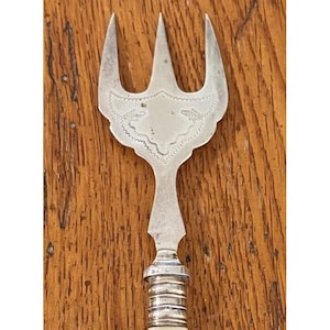 May include: Close-up of a silver fork with three tines. The fork has an ornate design on the handle, with a decorative shield-shaped element. The handle has a ribbed design near the base. The fork is set against a wooden background.