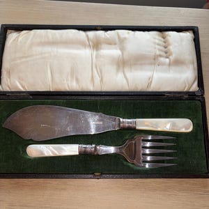 Puede incluir: Un juego de cubiertos para pescado plateados que consta de un cuchillo ancho y plano y un tenedor de cuatro dientes, ambos con mangos de nácar iridiscente y virolas de plata, presentados en un estuche rectangular con interior de terciopelo verde.
