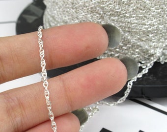 May include: Close-up of a silver-colored chain with a twisted rope design. The chain is held between two fingers, with a larger spool of the same chain visible in the background. The chain is likely used for jewelry making.