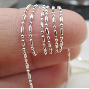Peut inclure: Gros plan sur une fine chaîne argentée avec un motif répétitif de petites perles rondes alternant avec des perles cylindriques allongées. La chaîne est tenue, mettant en valeur sa surface réfléchissante et son design complexe. Idéale pour la bijouterie.