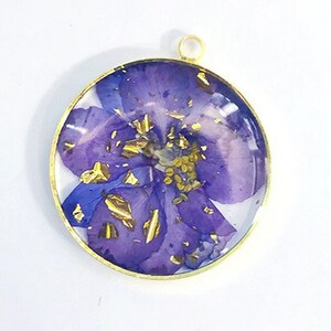 Puede incluir: Un colgante redondo de oro con una flor morada prensada en su interior. La flor tiene motas doradas por todas partes.