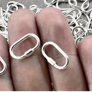 May include: Close-up of several small, shiny, silver-colored oval carabiner clasps. Three clasps are in focus, held between fingers, with a pile of similar clasps in the background. Each clasp has a hinged bar closure.