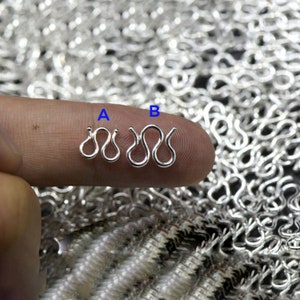 May include: Close-up of two silver-colored jewelry clasps, labeled A and B, held against a background of similar clasps. The clasps have a wavy design. The image is well-lit, highlighting the metallic sheen of the clasps.