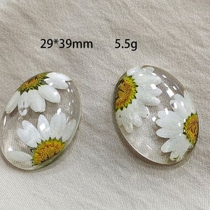 Peut inclure: Deux pendentifs ovales en résine transparente, chacun contenant une fleur de marguerite blanche avec un centre jaune. Les pendentifs mesurent 2,9 cm x 3,9 cm et pèsent 5,5 g. Fond neutre.
