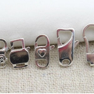 May include: Five silver-colored metal clasps of varying designs. The clasps include a lobster claw, a heart-shaped clasp, and a clasp with a decorative design. The clasps are displayed on a neutral background.