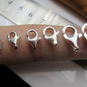 May include: A collection of six silver-colored lobster claw clasps of varying sizes. The clasps are arranged in a row, showcasing their different dimensions. The clasps are made of a shiny, reflective metal.