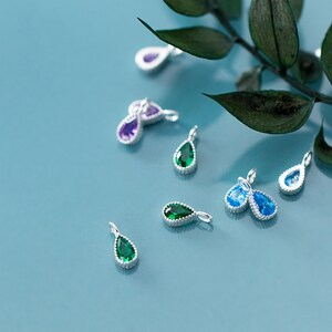 May include: A collection of teardrop-shaped pendants with silver settings and colorful gemstones. The pendants feature emerald green, amethyst purple, and light blue stones. The pendants are displayed on a light blue surface with green leaves.