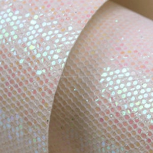 May include: Close-up of a roll of iridescent, textured fabric. The material has a light pink base with a grid pattern of small, shimmering, opalescent dots. The fabric is slightly curled, revealing its layered texture.