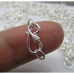 May include: A close-up of a silver-colored jewelry clasp, held between fingers. The clasp has a hook and loop design, with a textured center. Other clasps are visible in the background, suggesting a jewelry-making theme.