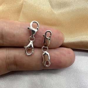 May include: Two silver-colored lobster claw clasps. The clasps are connected by a small ring. The clasps are designed to secure jewelry pieces. The background is a light gold color.