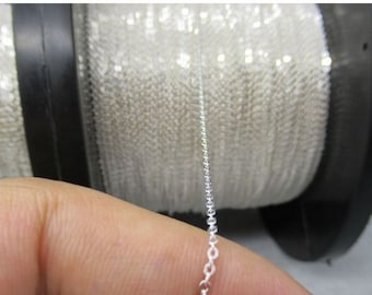 May include: Close-up of a silver chain held in a person's hand. The chain is delicate and has small, round links. A large spool of chain is visible in the background.