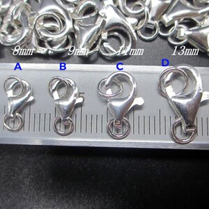 May include: Close-up of several silver-colored lobster claw clasps of varying sizes, ranging from 8mm to 14mm. The clasps are arranged on a ruler for size comparison. The image shows the clasps' hook and ring details.