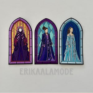 May include: Three embroidered patches in the shape of stained glass windows. Each patch features a figure in a long gown. The first patch has a figure in purple and gold, the second in purple, and the third in blue. The text "ERIKAALAMODE" is at the bottom.