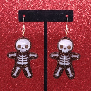 May include: A pair of skeleton gingerbread man earrings with white and brown glitter. The earrings are on a black earring stand against a red glitter background.