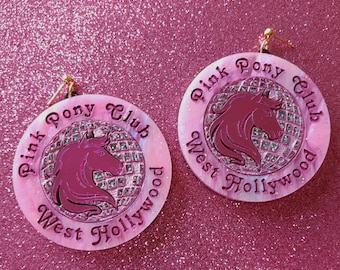 May include: A pair of pink and white round earrings with a pink horse silhouette and the text "Pink Pony Club West Hollywood" on each earring.
