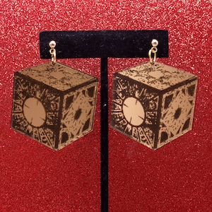 May include: A pair of gold-toned earrings shaped like a cube with intricate details. The earrings are hanging from a black display stand.