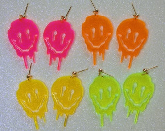 May include: Six pairs of neon-colored acrylic earrings. Each earring is shaped like a dripping smiley face. The earrings are available in pink, yellow, and green.