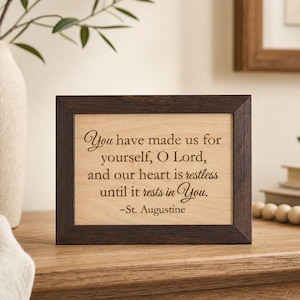 May include: A rectangular wooden sign with a dark brown frame and a light tan center panel. The text reads: You have made us for yourself, O Lord, and our heart is restless until it rests in You. -St. Augustine, engraved in a dark brown font.