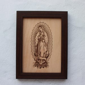 May include: A framed wood carving of Our Lady of Guadalupe, a religious icon, in a dark brown frame. The carving is on a light wood background.