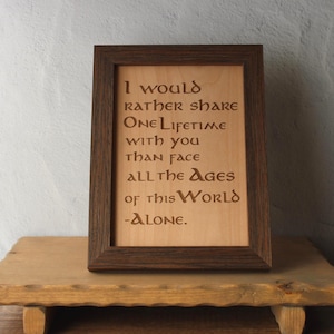 Op de afbeelding: Houten lijst met de tekst "Ik zou liever één leven met je delen dan alle tijden van deze wereld -Alleen- onder ogen zien." De lijst is donkerbruin en de tekst is gegraveerd op een licht houten oppervlak.