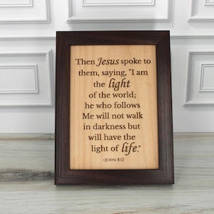 May include: A framed wooden plaque with a dark brown frame. The plaque features engraved text from John 8:12, including the words "I am the light of the world." The background is a light wood grain.