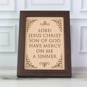 Può includere: Targa in legno incorniciata con la scritta "LORD JESUS CHRIST SON OF GOD HAVE MERCY ON ME A SINNER" in una cornice marrone scuro. La targa presenta decorazioni angolari e uno sfondo chiaro con venature del legno.