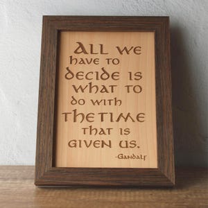 Könnte beinhalten: Holzgerahmtes Kunstwerk mit dem Zitat "All we have to decide is what to do with the time that is given us." Der Text ist in das helle Holz geschnitzt, umrahmt von einem dunkelbraunen Holzrahmen.