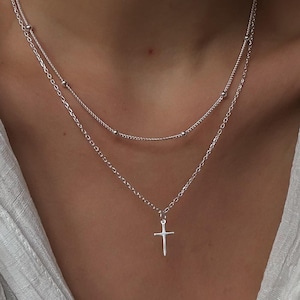 May include: A silver chain necklace with a small silver cross pendant. The necklace is layered with a second, thinner chain.