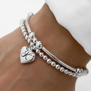 May include: A silver charm bracelet with a heart charm engraved with the letter "K" and a silver beaded bracelet.